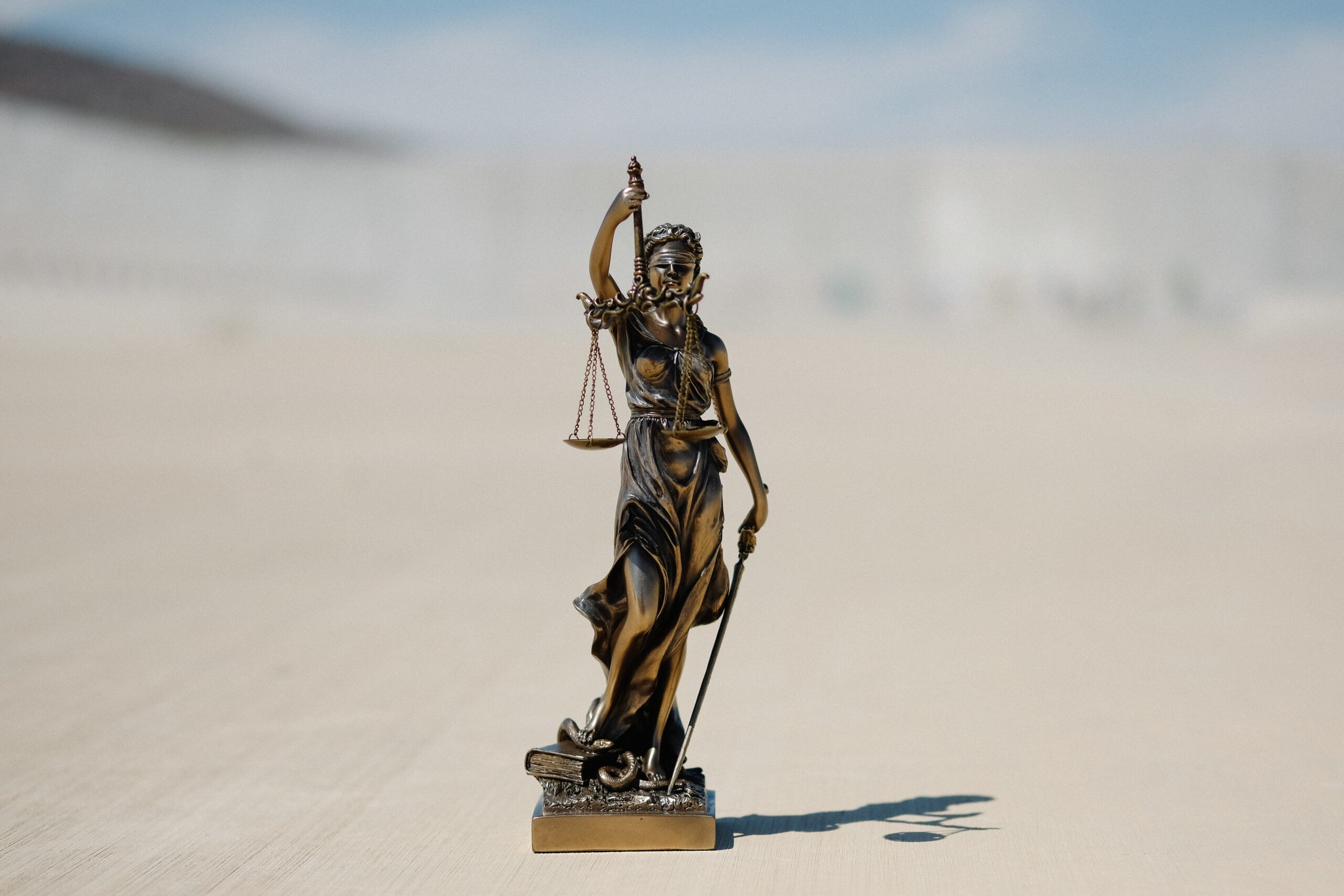 Justitia mit der Waage in der einen Hand und dem Schwert zum Boden gerichtet in der anderen. Sie steht auf einem Sockel, auf dem u. a. ein Buch liegt.